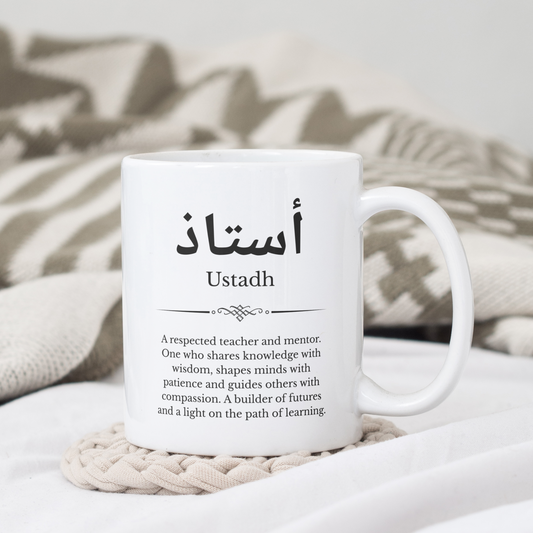 White mug with Arabic and English word “Ustadh” and its meaning – placed on beige coaster with neutral blanket, Islamic teacher appreciation gift.