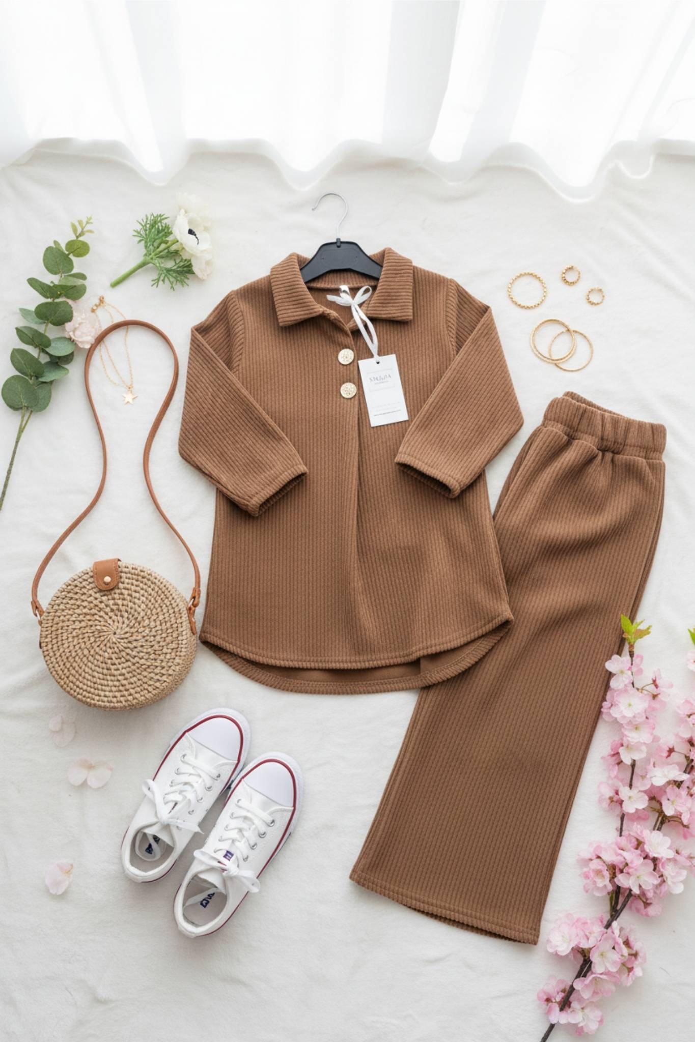 Brown outfit with shirt, pants, and shoes on a white surface with flowers and a bag.